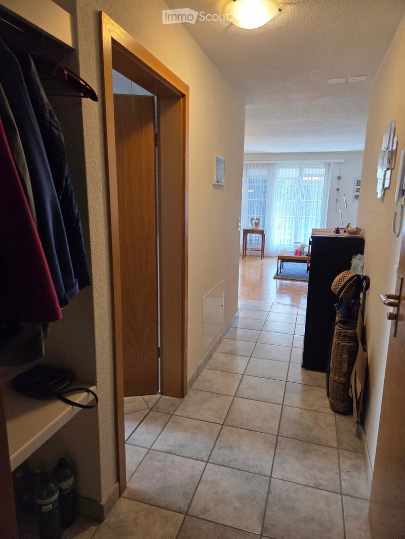 A narrow hallway with tiled flooring, leading to an open living area. Visible are a coat rack with jackets, a small table, and a wicker basket in the background.