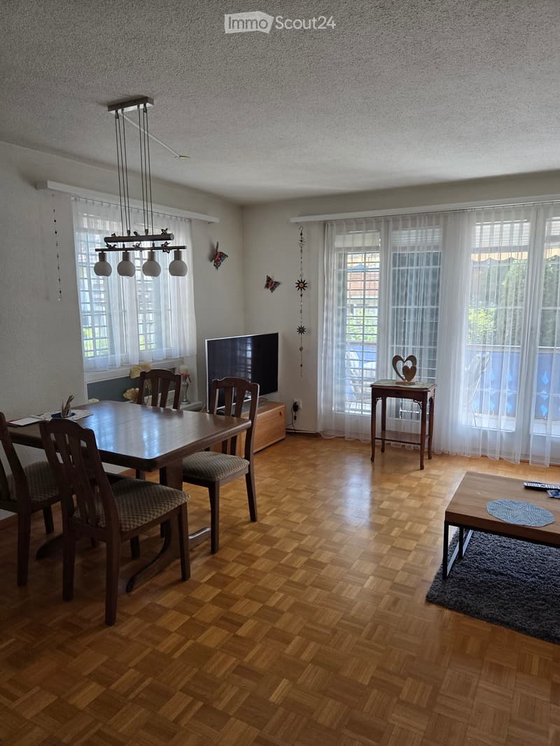 Dining and living area with wooden flooring, large windows with white curtains, a dining table with chairs, a television, a side table, and a floor lamp.