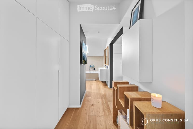Modern hallway with light wooden flooring, white walls, built-in white cabinets, and a small candle on a wooden shelf. A bathroom is visible at the end of the hallway.