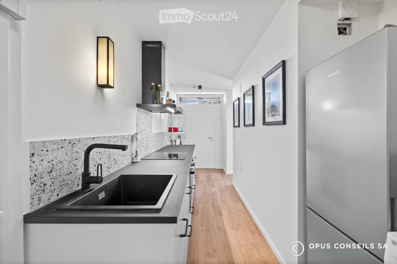 Modern kitchen with white cabinets, a black sink, a stainless steel faucet, a refrigerator, and a shelf with bottles. The floor is light wood, and there are framed pictures on the wall.