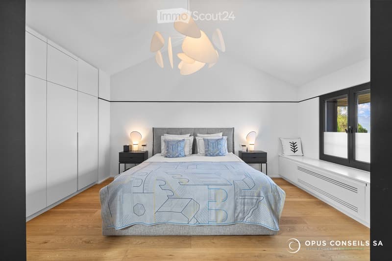 Modern bedroom with a large bed, nightstands, lamps, and a window with a view. The room features a contemporary design with white walls, wooden flooring, and built-in storage.
