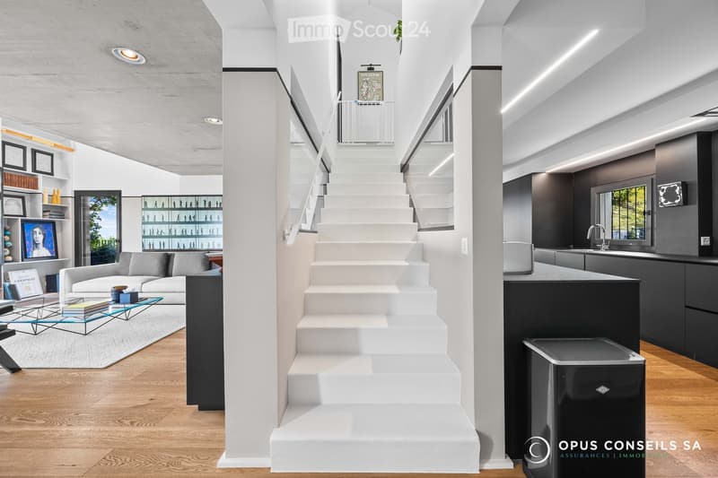 Modern, open-plan living space with a grey sofa, glass coffee table, books, framed artwork, shelves, a staircase leading to an upper level, and a sleek kitchen area with a counter, sink, and trash bin.