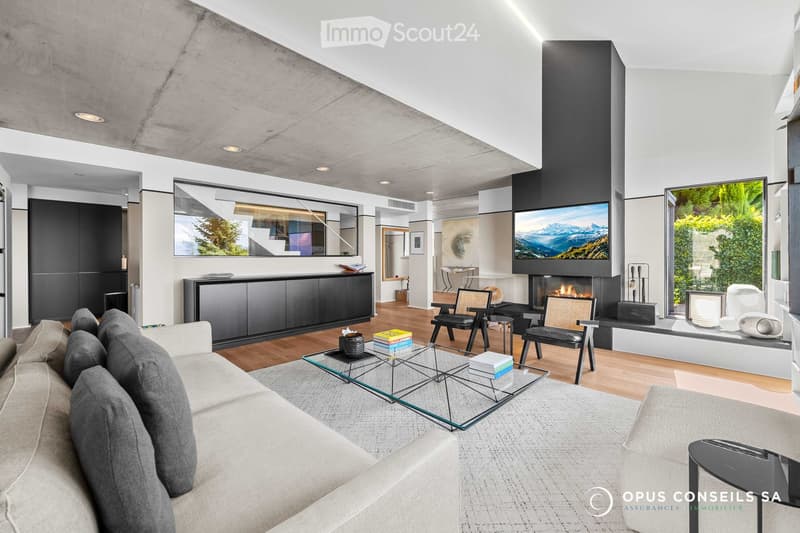 Modern living room with a fireplace, large TV, sofa, coffee table, armchairs, and a view of greenery through large windows.