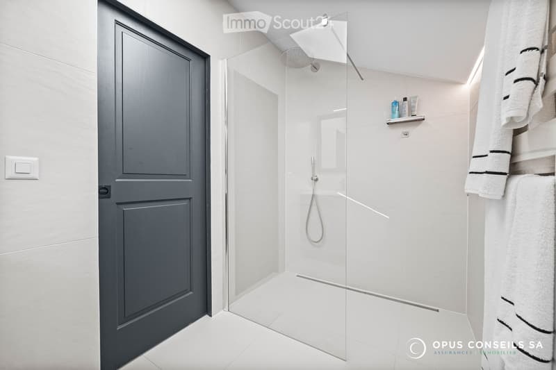 Modern bathroom with a glass-enclosed shower, white tiles, a towel rack, and a small shelf holding toiletries.