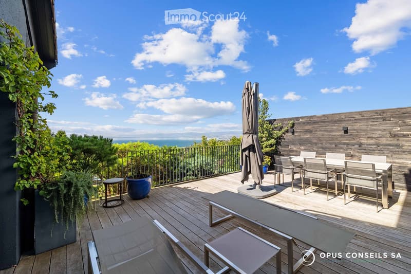 Wooden terrace with outdoor furniture, including a dining table, chairs, a lounge chair, and an umbrella, overlooking a scenic view.
