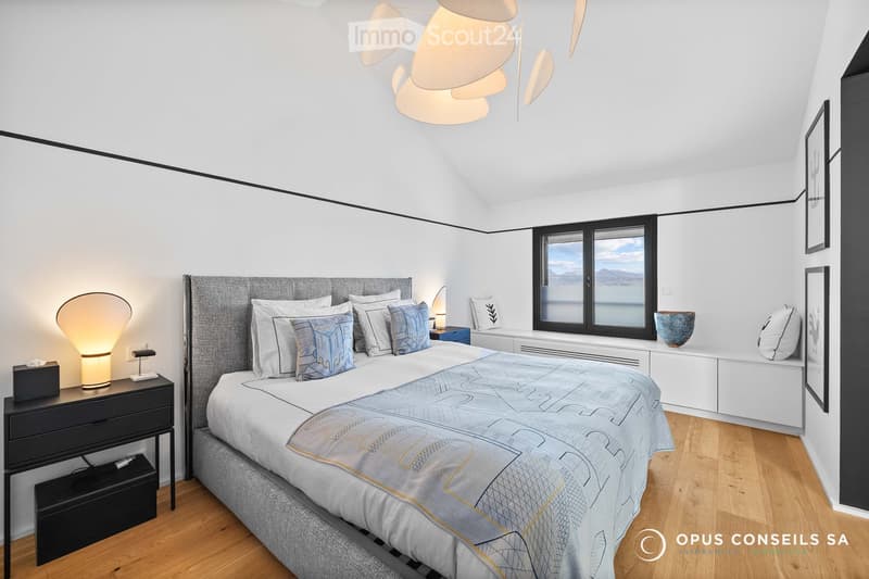 Modern bedroom with a large bed, nightstand, lamp, and a view of the lake through large windows.