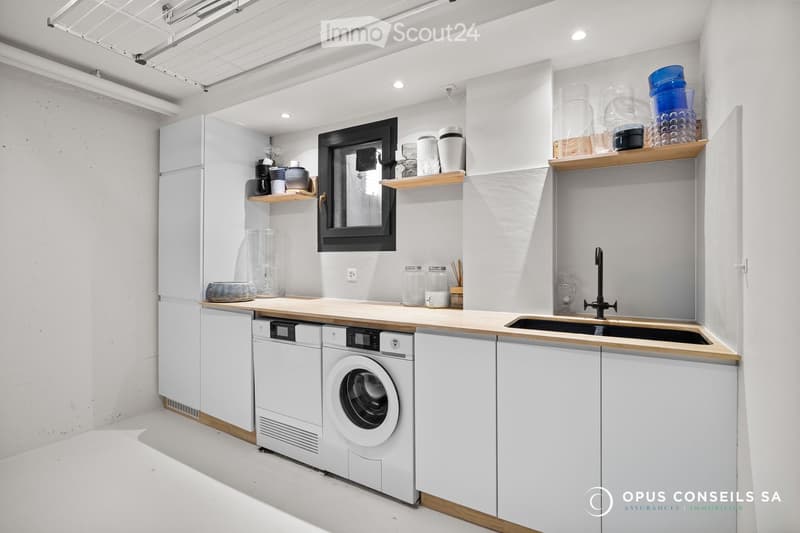 Modern kitchen with white cabinets, wooden countertops, stainless steel sink, and built-in appliances including a refrigerator, washing machine, and dishwasher. Shelves hold cups, jars, and other kitchen items.
