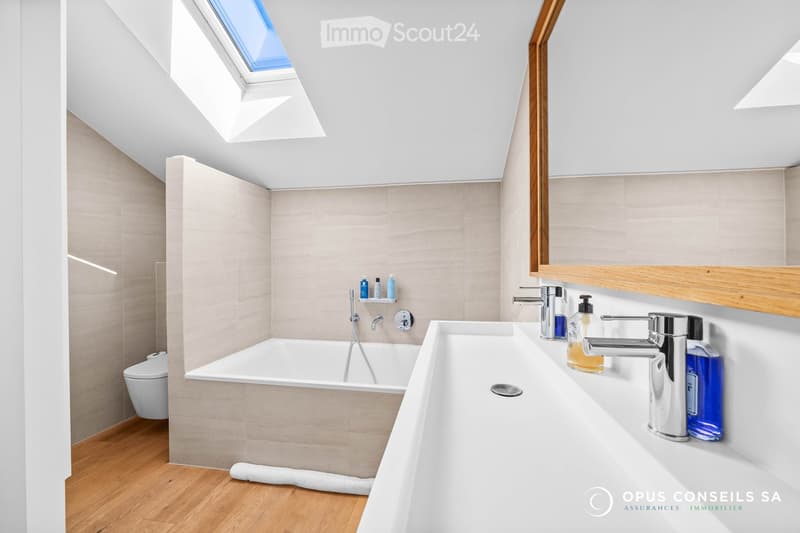 Modern bathroom with a bathtub, toilet, sink, and mirror. Features a skylight window and wooden flooring.