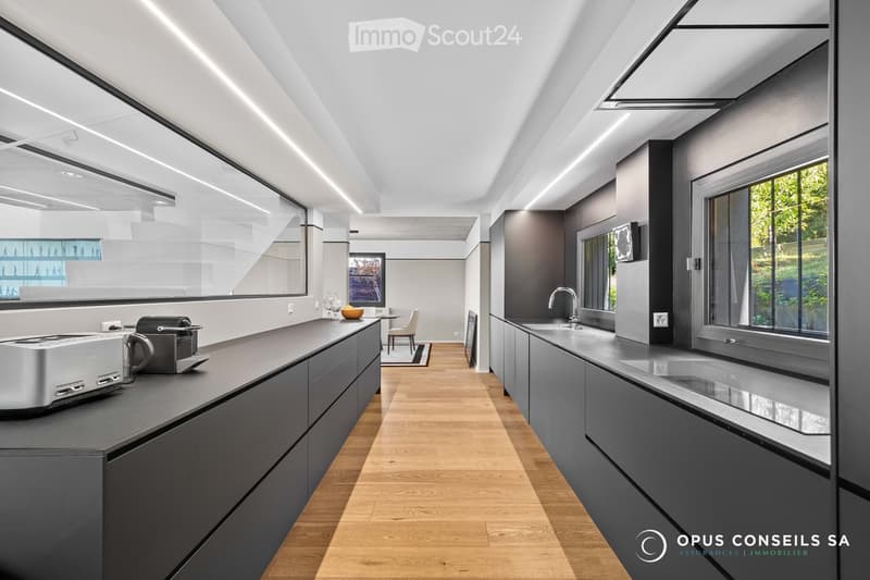 Modern kitchen with dark grey cabinets, wooden flooring, and large windows. Features a toaster, coffee machine, sink, and a small dining area with a chair and table.