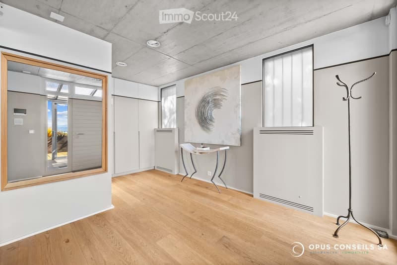 Modern, minimalist room with light wooden flooring, white walls, and contemporary decor including a small table, chair, coat rack, and a large mirror. The room features built-in storage units and a radiator.