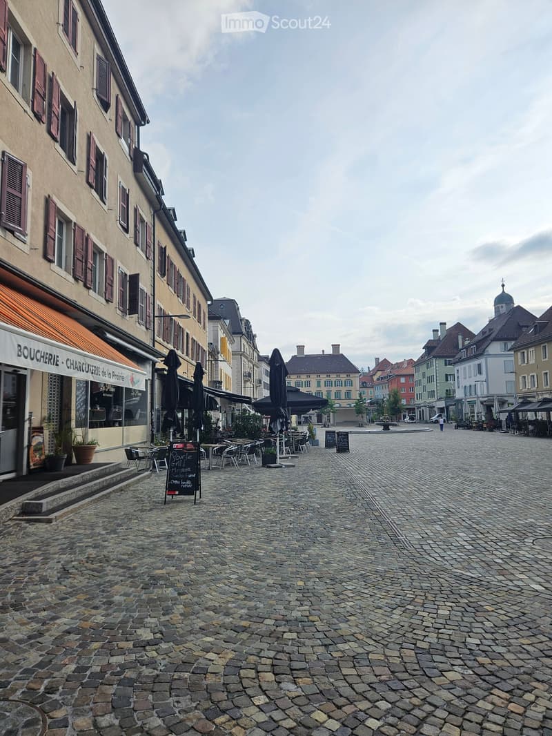 A cobblestone square with outdoor seating, closed umbrellas, and buildings with shutters and awnings.