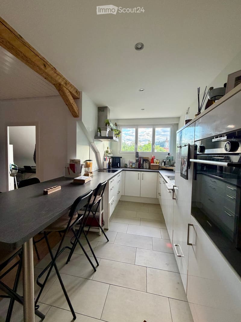 Modern kitchen with white cabinets, a dark countertop, and a dining area with a table and chairs. Appliances include a refrigerator, oven, microwave, and coffee maker. There is a window with a view of greenery and a tower.