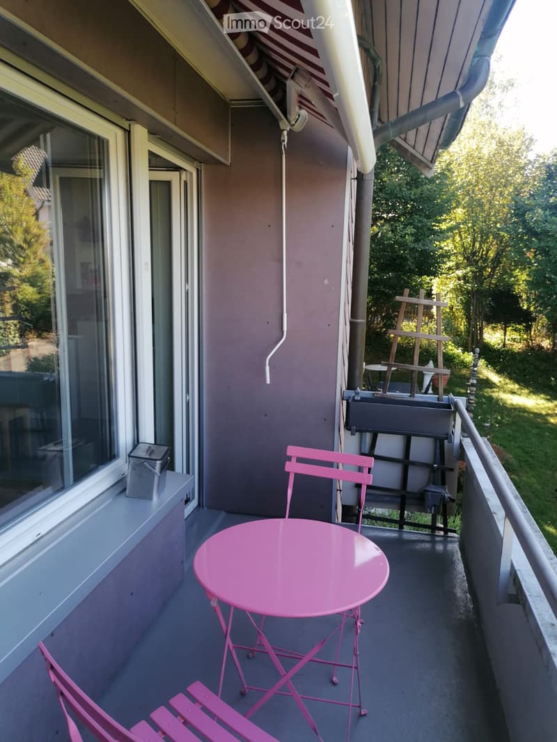 Balcony with pink metal table and chairs, overlooking a green area with trees.