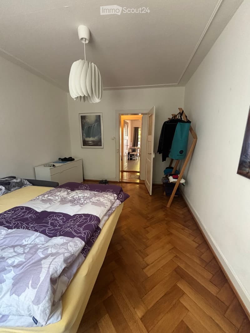 Bedroom with wooden flooring, a bed with a purple and white bedspread, a nightstand, a picture frame on the wall, and an open doorway leading to another room with a chair and table visible.