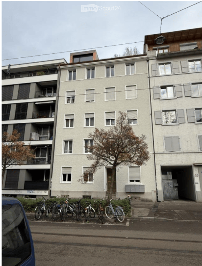 Multi-story residential building with balconies, bicycles parked in front, and a small tree near the entrance.