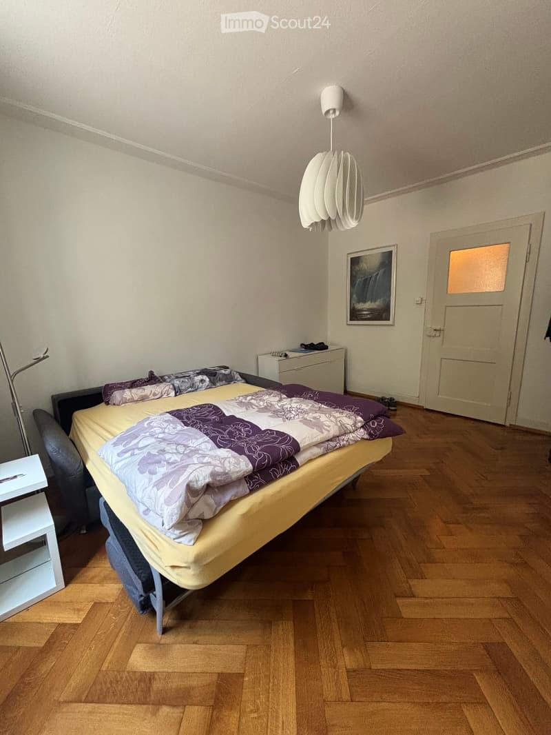 Cozy bedroom with wooden flooring, a neatly made bed with a colorful quilt, a small table with a lamp, a chair, and a picture frame on the wall.