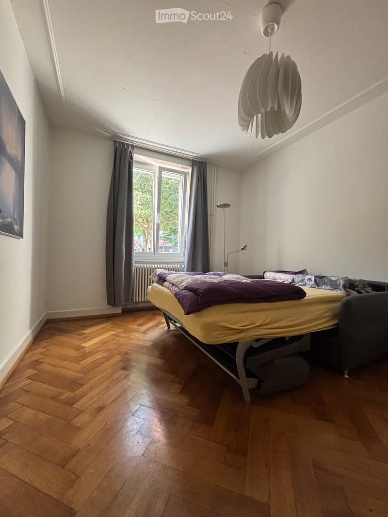 Bright bedroom with wooden herringbone flooring, large window with grey curtains, bed with yellow and purple bedding, radiator, picture frame, and a hanging lamp.