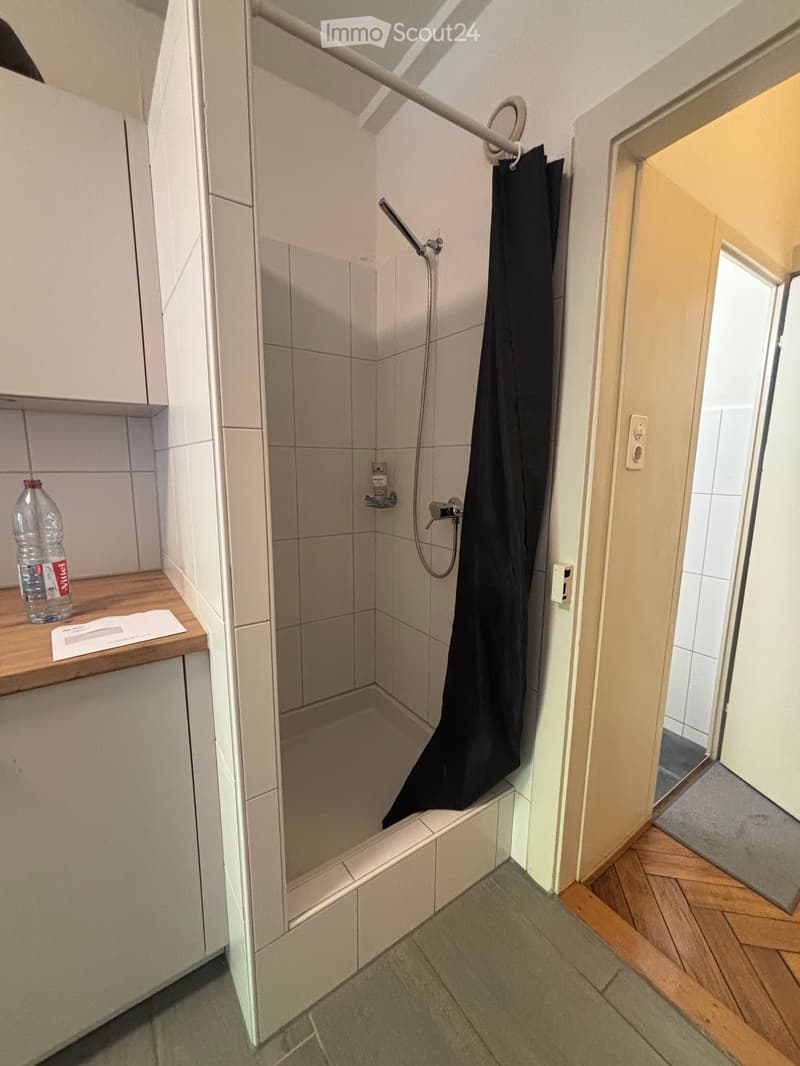 Small bathroom with tiled walls, a shower area with a curtain, a wooden shelf holding a bottle, and a cabinet.