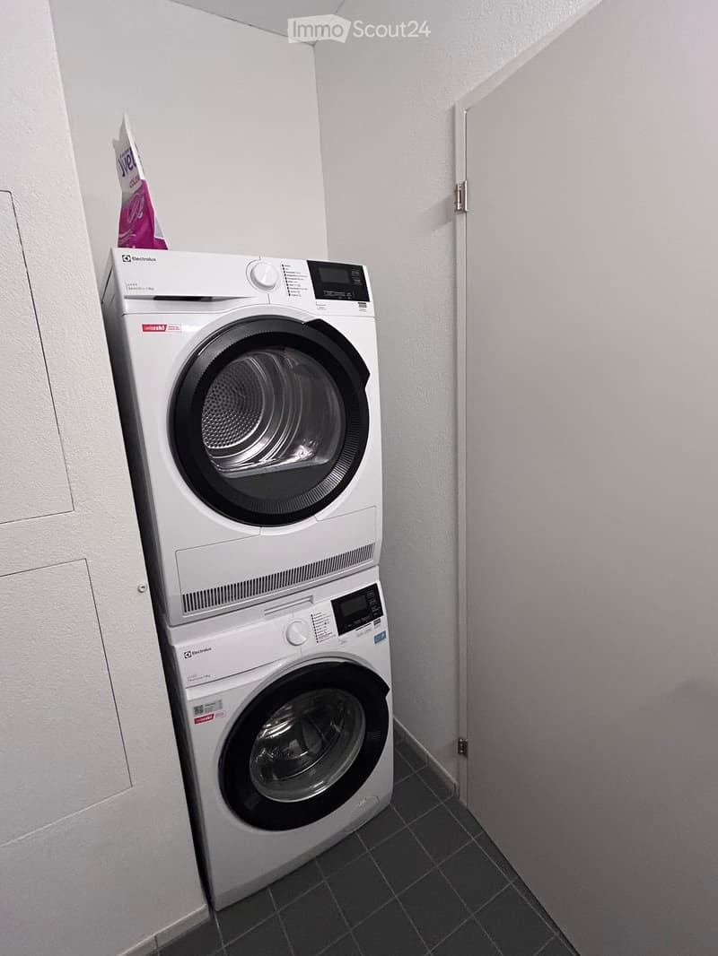 A small bathroom with a white washing machine and dryer stacked vertically, white tiled walls, and a dark tiled floor.