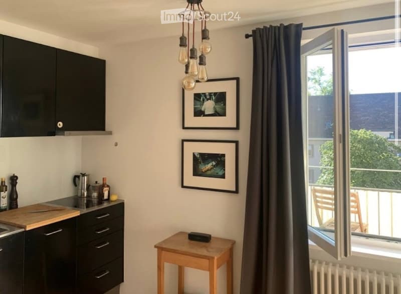 Modern kitchen with black cabinets, a wooden cutting board, a small wooden stool, and two picture frames on the wall. A window with a curtain opens to a balcony.