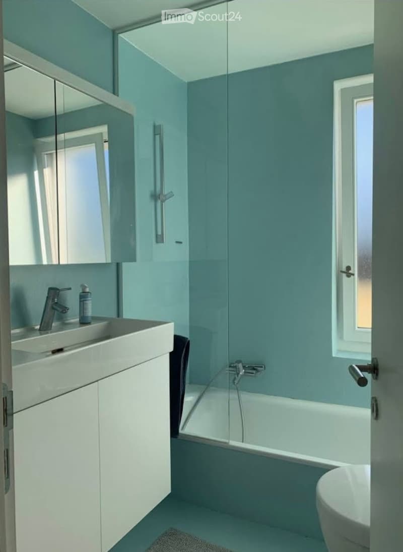 Modern bathroom with light blue walls, white fixtures, a glass-enclosed bathtub, a sink with a modern faucet, and a mirror above the sink.