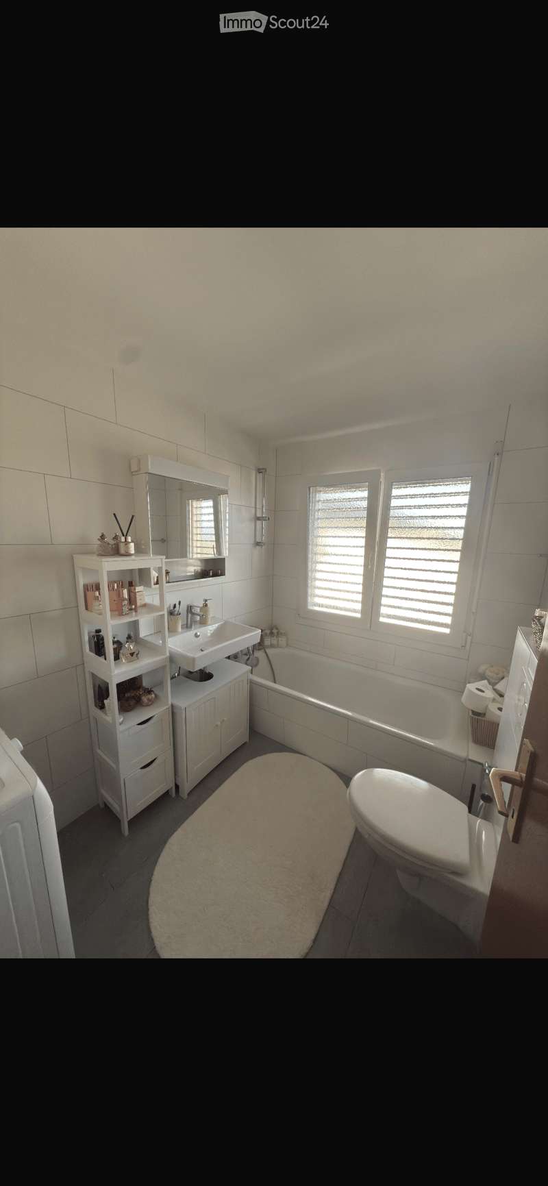 Modern bathroom with white tiles, a bathtub, toilet, sink, mirror, and a white bathroom cabinet with toiletries. The room features a large window with blinds and a plush beige rug on the floor.