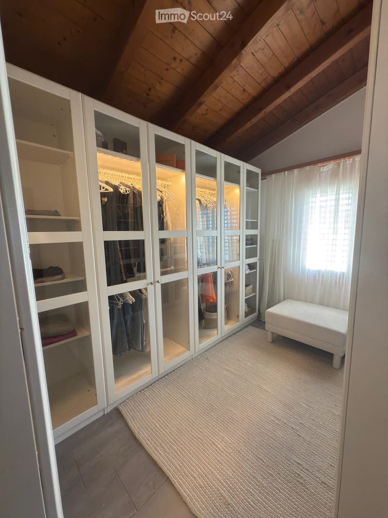 Attic room with wooden slanted ceilings, white wardrobe with mirrored doors, and a small white stool on a textured rug.