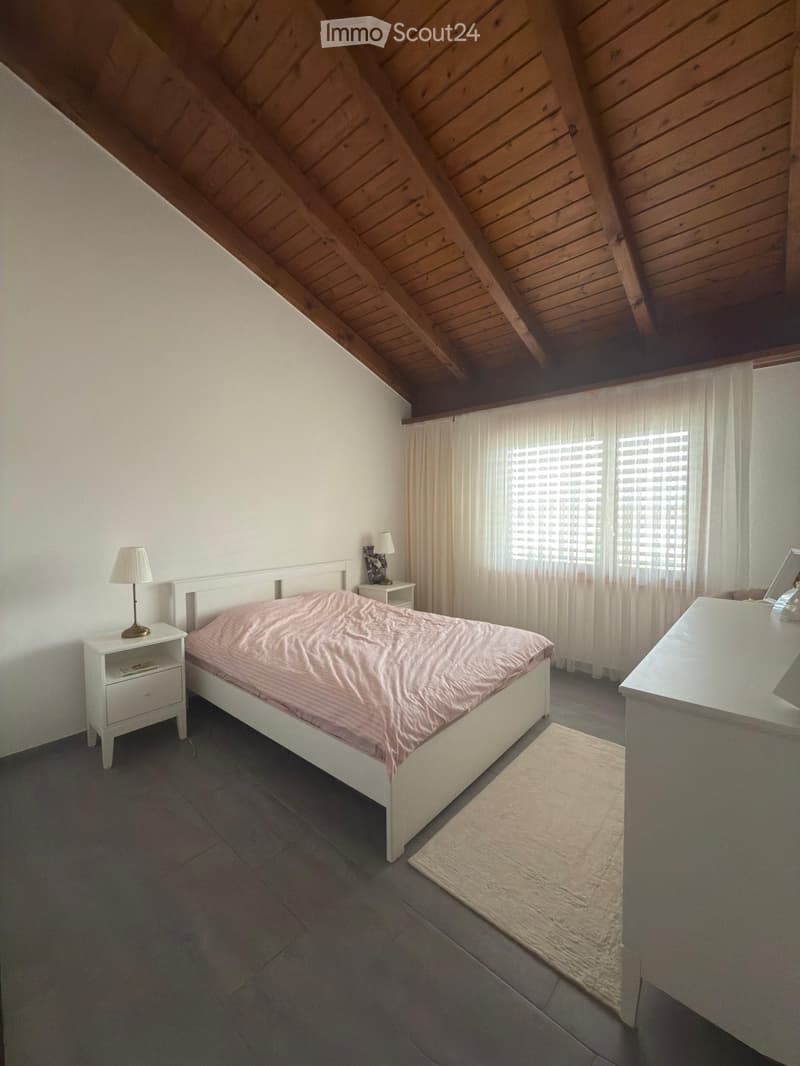 Cozy attic bedroom with wooden slanted ceilings, grey tiled floor, white walls, and large windows with white curtains. Features a white bed with pink bedding, two nightstands with lamps, a small rug, and a mirror on the dresser.