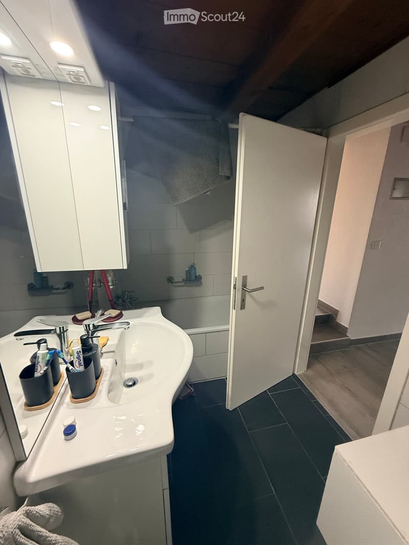 Modern bathroom with a white sink, mirror cabinet, toothbrush holders, and a towel on the floor. The shower area is visible through an open door.