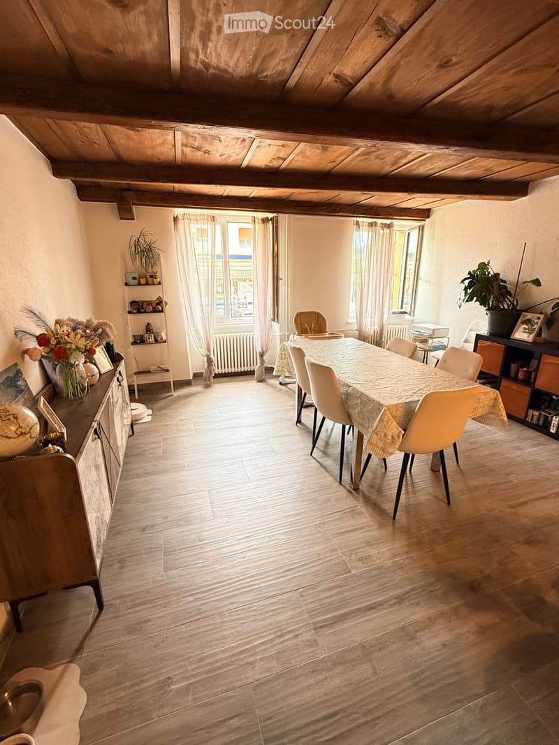 Bright dining area with wooden floors, large windows, a dining table with chairs, a cabinet with decorative items, and shelves with plants and books.
