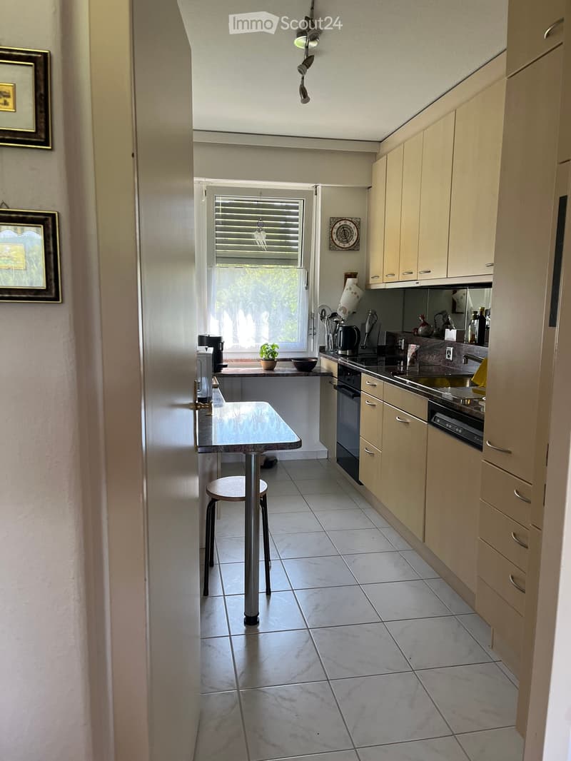 Modern kitchen with light-colored cabinets, tiled floor, and a central island with a table and stool. Appliances include a fridge, oven, dishwasher, and coffee maker. A small plant and a bowl are on the island.