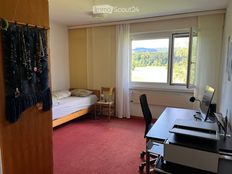 Cozy bedroom with a single bed, desk, chair, and large windows offering a scenic view. The room features a red carpet, a nightstand with a lamp, and a curtained window.