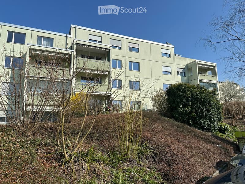 Modern multi-story residential building with light green facade, multiple windows, and balconies. The building is surrounded by sparse vegetation and a raised ground floor.