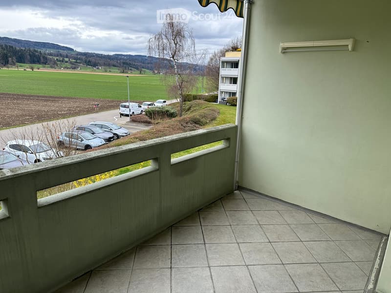 Balcony with tiled floor, white railing, overlooking a parking area, green fields, and distant hills.