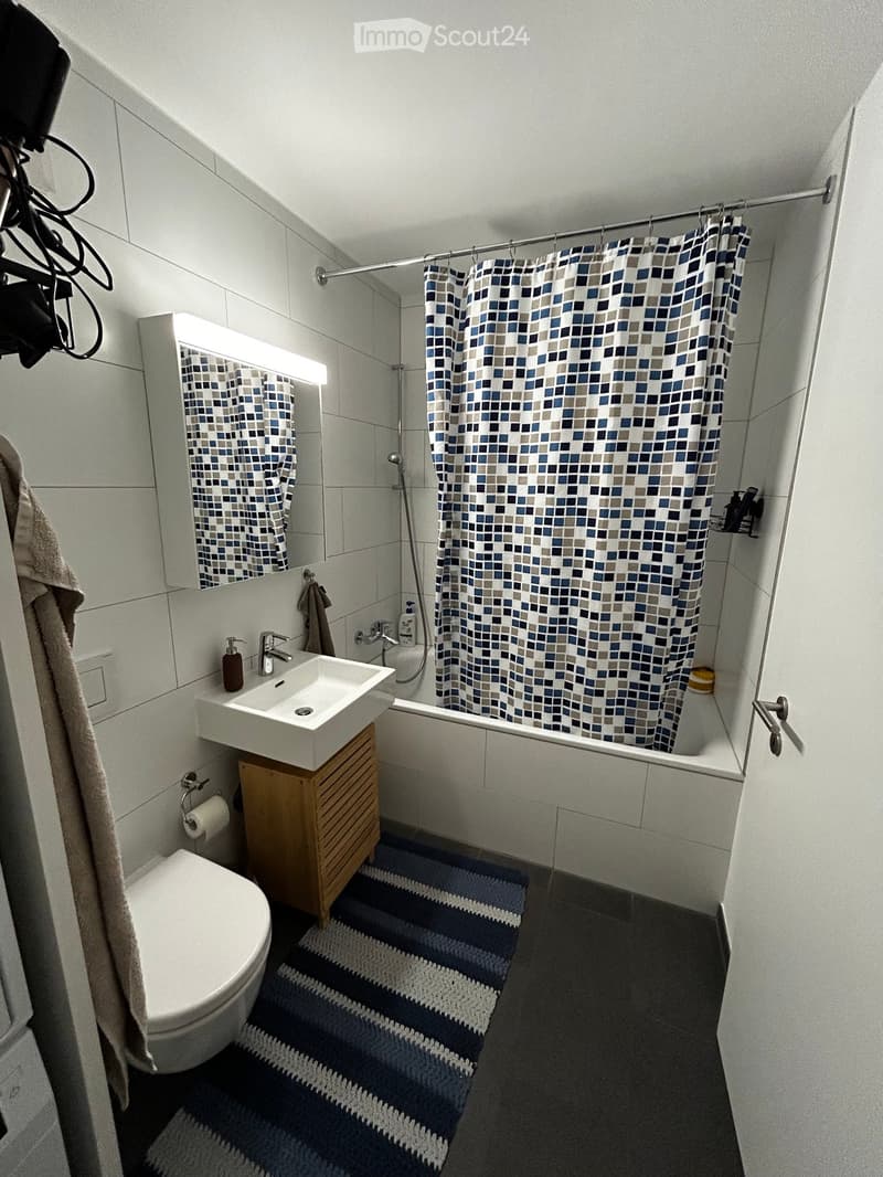 Modern bathroom with white tiles, a white sink, a toilet, a bathtub with a blue and white checkered shower curtain, a large mirror with built-in lighting, and a blue and white striped rug.
