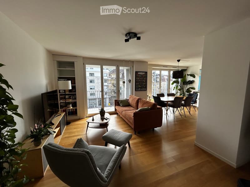 Modern living room with wooden flooring, large windows, balcony access, sofa, coffee table, ottoman, dining area with table and chairs, television, and decorative plants.