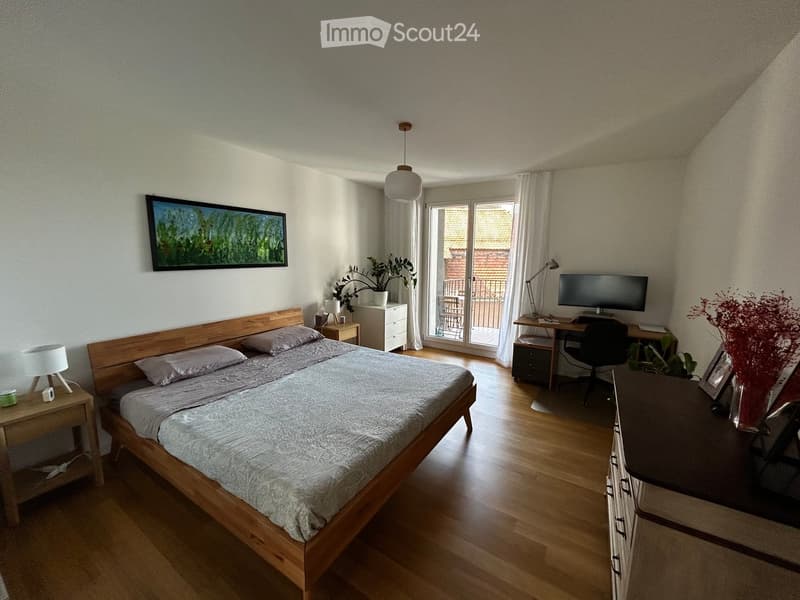 Bright bedroom with wooden floor, double bed, nightstand, desk with chair, and large window leading to a balcony.