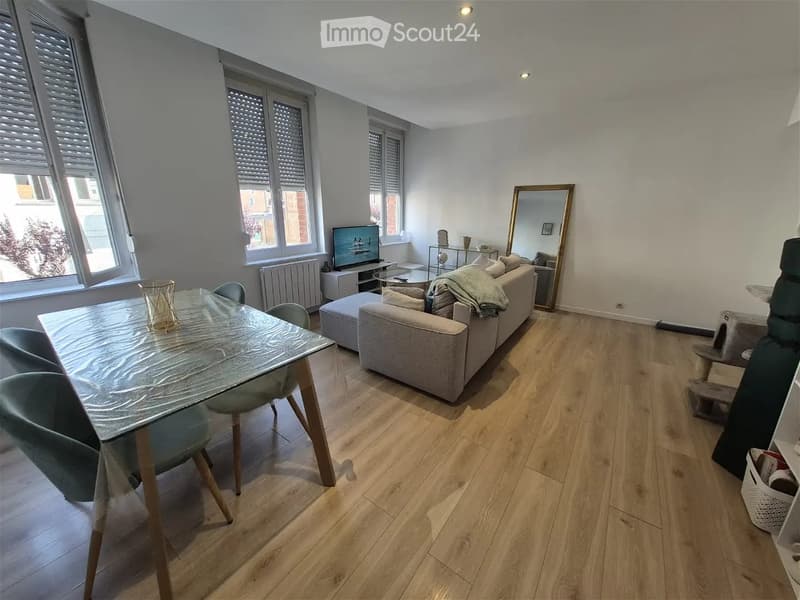 Modern living room with light wooden flooring, large windows, a grey sofa, a glass dining table with chairs, a TV stand, and a decorative mirror.