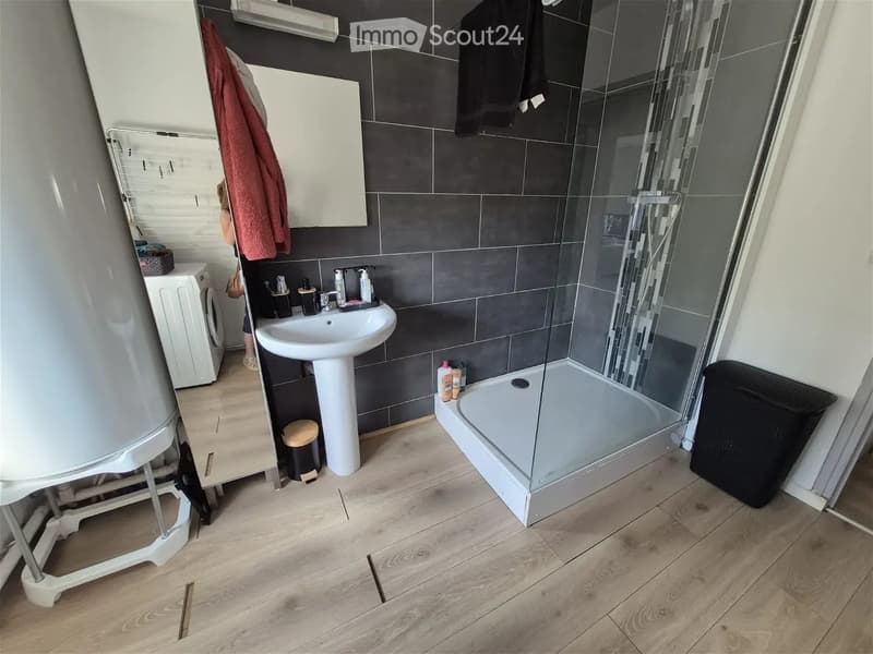 Modern bathroom with dark grey tiles, white sink, glass-enclosed shower, washing machine, and wooden flooring.