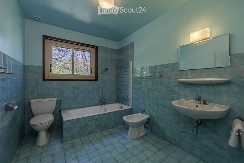 Bathroom with a toilet, bathtub, bidet, sink, mirror, and towel. The room features light blue tiles and a window with a view of trees.