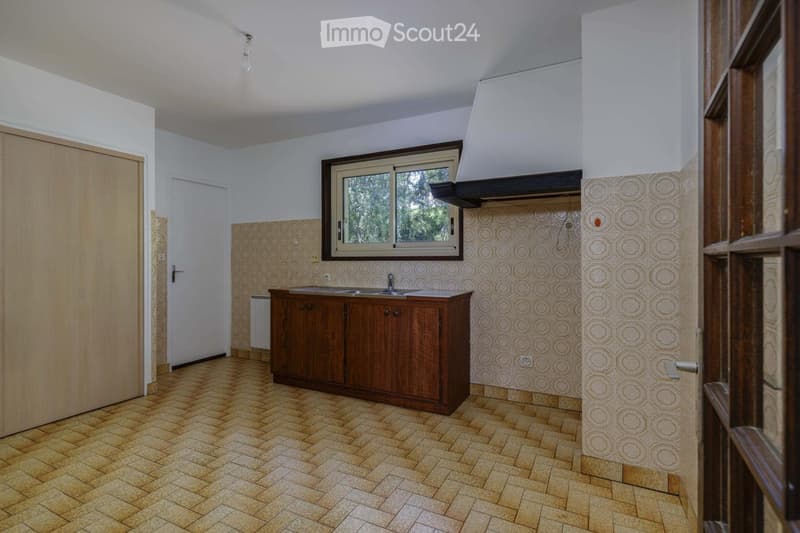 Empty kitchen with wooden cabinets, a sink, and a window above it. The walls are tiled with a circular pattern, and the floor is made of herringbone-patterned tiles.