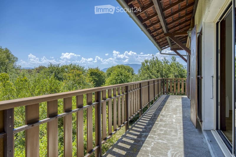 Balcony with wooden railing, overlooking a lush green landscape and distant mountains under a clear blue sky.