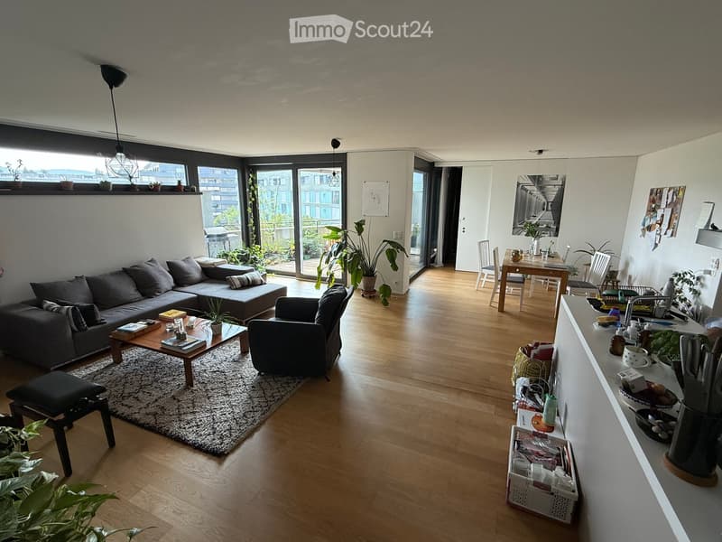 Open-plan living and dining area with wooden flooring, large windows, and a balcony. Features a grey sectional sofa, coffee table, armchair, dining table with chairs, and kitchen counter with various items.
