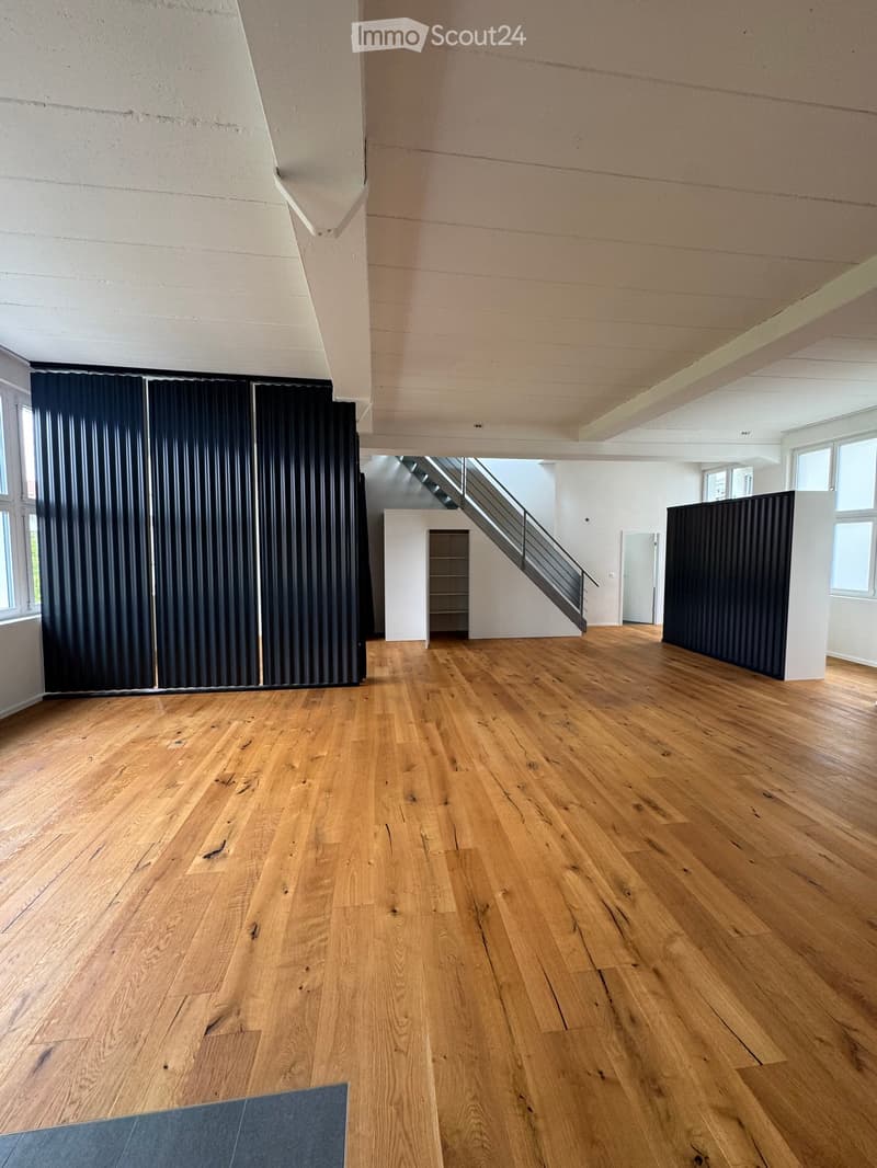 Empty room with wooden flooring, white walls, and large windows. Features a staircase leading to another level and dark paneling on one wall.
