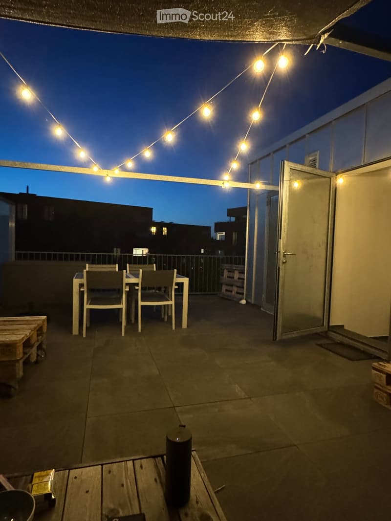 Balcony with a small table and chairs, string lights, and a view of the city at dusk.