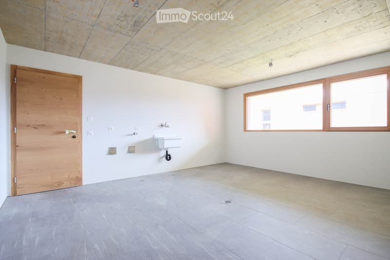 Empty room with white walls, grey tiled floor, wooden door, and a small sink with a faucet. The ceiling features a mix of exposed stone and wooden panels, and there are two large windows with wooden frames allowing natural light to enter.