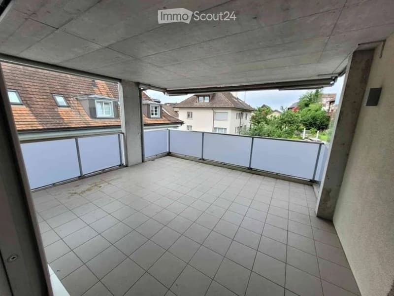 Empty balcony with tiled floor and white railing, overlooking neighboring houses and greenery.
