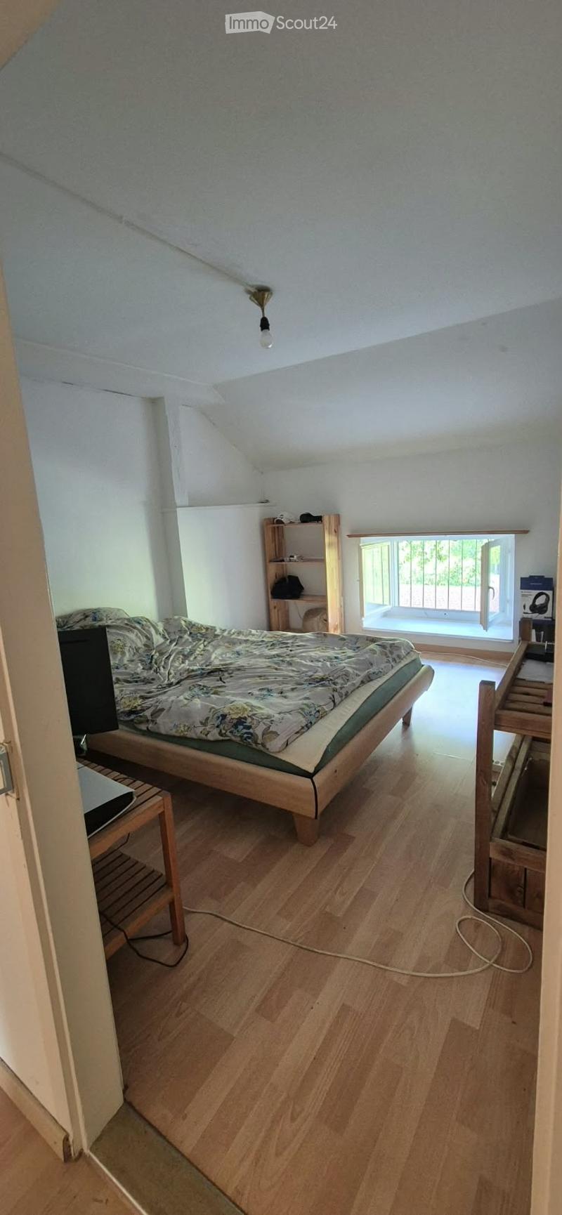 Bright bedroom with wooden flooring, slanted ceiling, and large window with balcony access. Features a wooden bed with patterned bedding, a nightstand, a small shelf, and a laptop on a wooden stand.