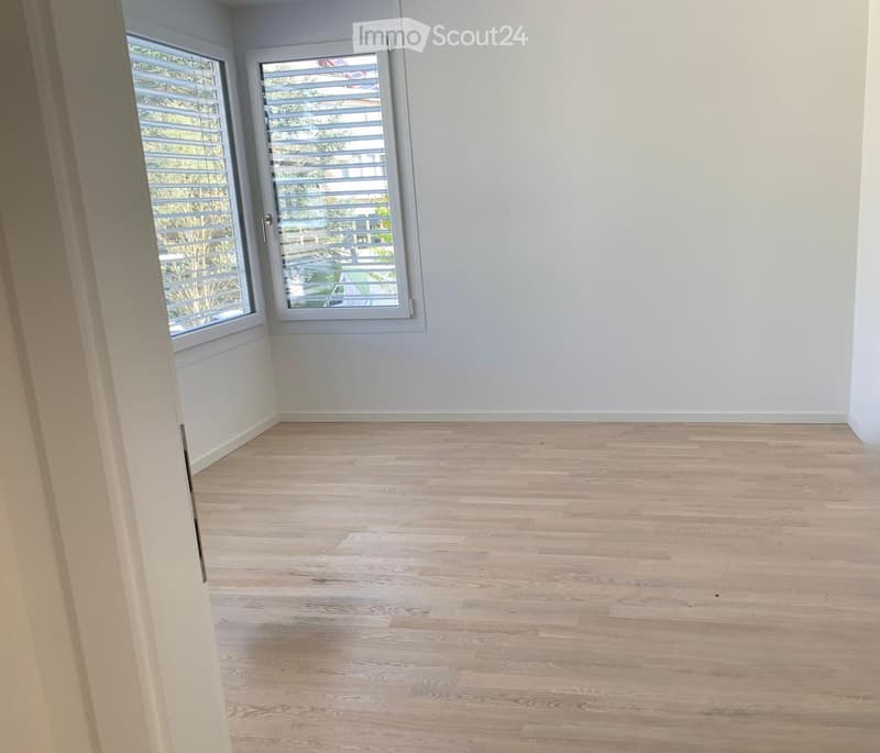 Empty room with light wooden flooring and white walls, featuring large windows with blinds and a door leading outside.