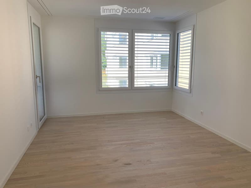 Empty room with light wooden flooring and white walls, featuring large windows with blinds and a glass door.